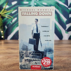 May include: A VHS tape of the movie Falling Down starring Michael Douglas. The cover features a man in a white shirt and tie holding a briefcase and a tool, with the text The adventures of an ordinary man at war with the everyday world.