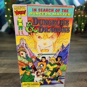 May include: A Dungeons & Dragons VHS tape titled In Search of the Dungeon Master. The cover features a large, menacing face above a group of animated characters, including a knight, a wizard, and a unicorn, with the text The Forces of Good Fight for Supremacy Over Evil.