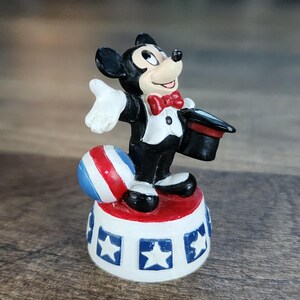 May include: A Mickey Mouse figurine in a magician's outfit, standing on a white and red platform with blue stars. The figurine has a red bow tie, a black top hat, and a red, white, and blue ball.