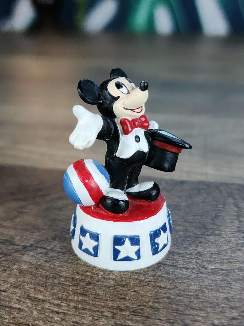May include: A Mickey Mouse figurine in a magician's outfit, standing on a white and red platform with blue stars. The figurine has a red bow tie, a black top hat, and a red, white, and blue ball.