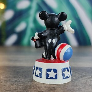May include: A small figurine of a black mouse in a tuxedo, white gloves, and a red hat, standing on a white base with blue stars. A red, white, and blue ball is next to the mouse. The figurine is a circus-themed collectible.