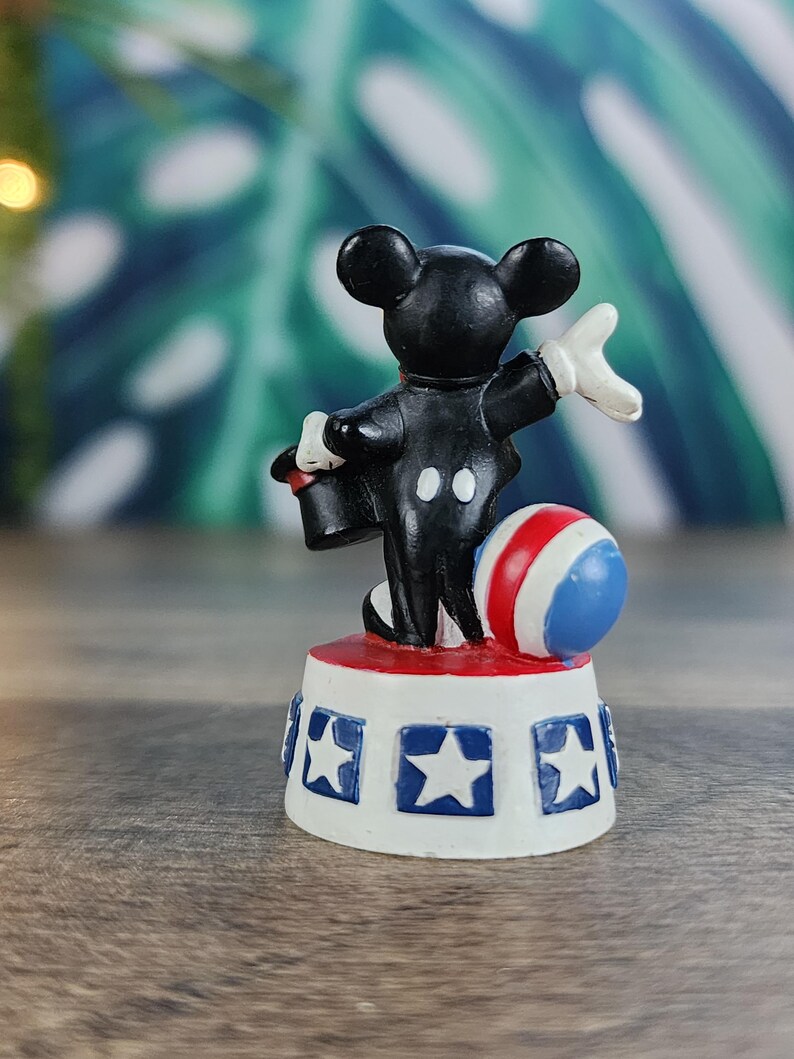 May include: A small figurine of a black mouse in a tuxedo, white gloves, and a red hat, standing on a white base with blue stars. A red, white, and blue ball is next to the mouse. The figurine is a circus-themed collectible.
