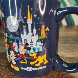 May include: A navy blue ceramic mug featuring a colorful illustration of Disney characters and landmarks. The mug includes Mickey Mouse, Minnie Mouse, Pluto, and a castle. The words "Walt Disney World" are printed on the mug.