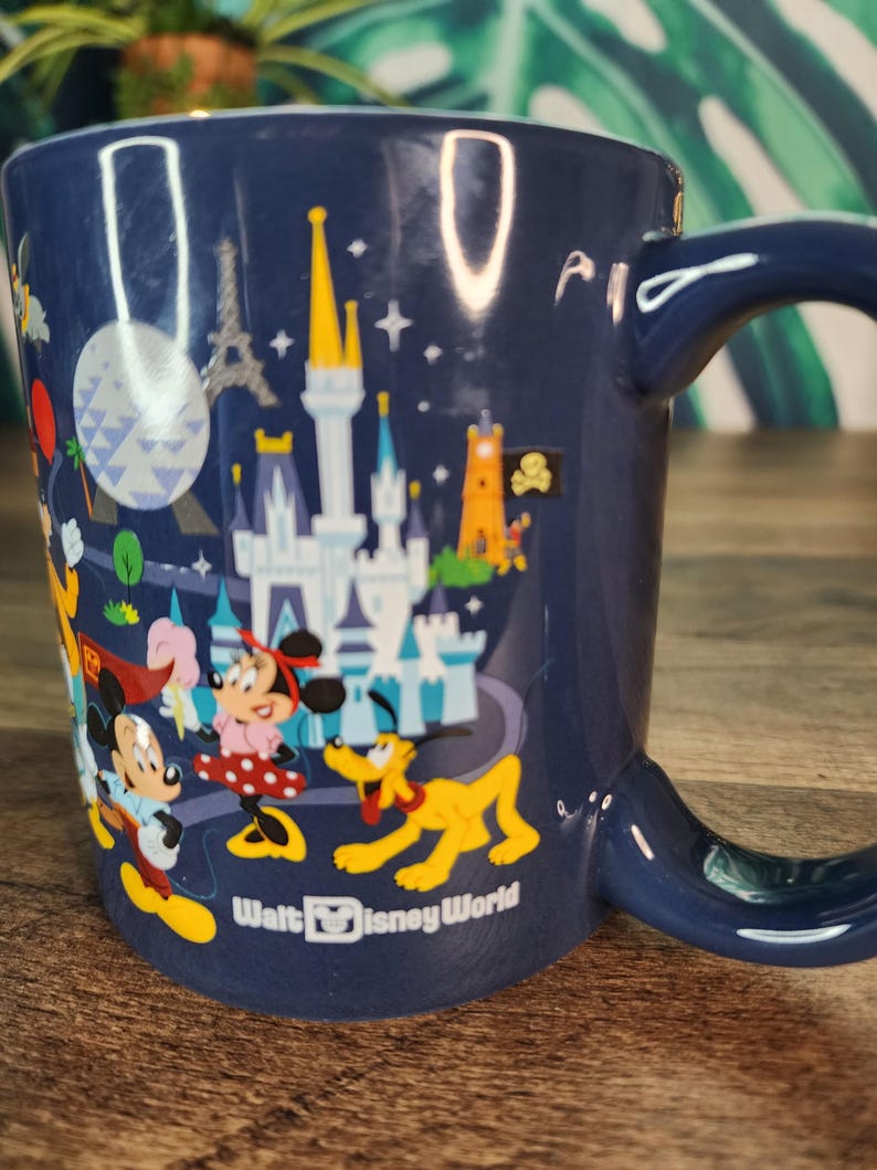 May include: A navy blue ceramic mug featuring a colorful illustration of Disney characters and landmarks. The mug includes Mickey Mouse, Minnie Mouse, Pluto, and a castle. The words "Walt Disney World" are printed on the mug.