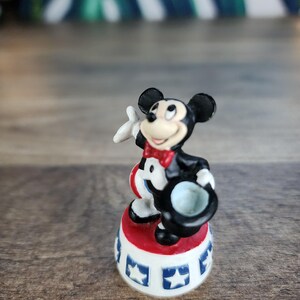 May include: A small Mickey Mouse figurine in a circus ringmaster outfit. The figure is standing on a red and white base with blue star accents. Mickey is wearing a black tuxedo, red bow tie, and holding a black top hat.
