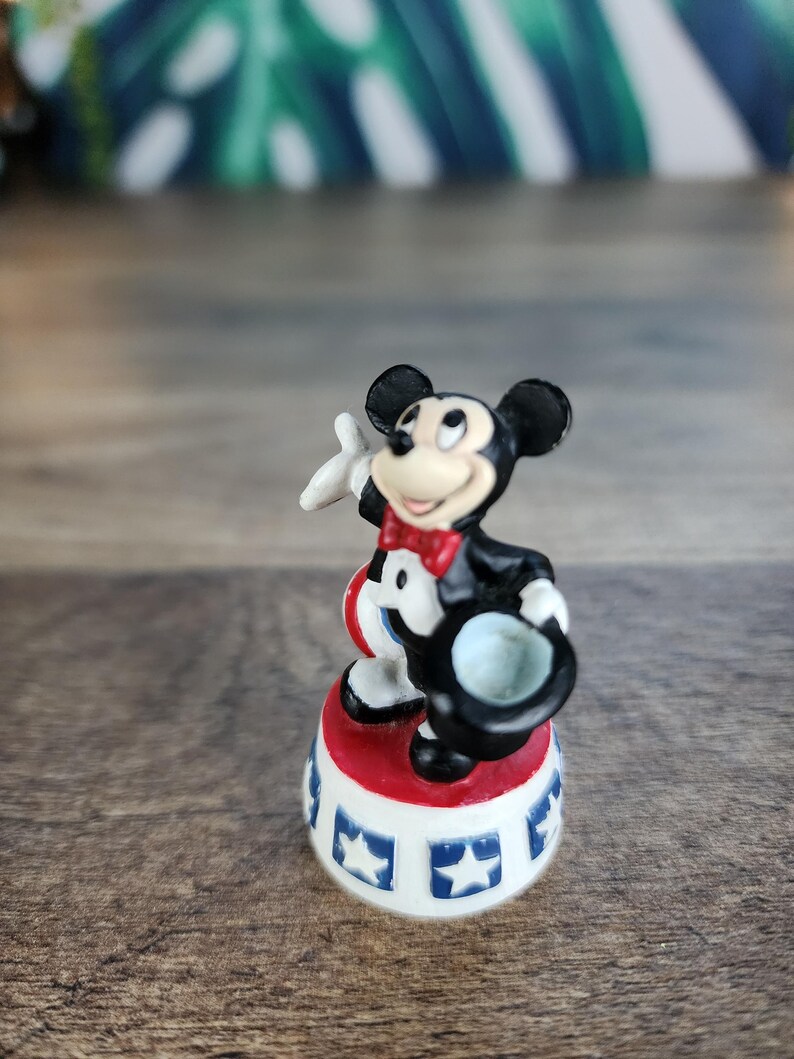 May include: A small Mickey Mouse figurine in a circus ringmaster outfit. The figure is standing on a red and white base with blue star accents. Mickey is wearing a black tuxedo, red bow tie, and holding a black top hat.