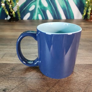 May include: A blue ceramic mug with a light blue interior. The mug has a large handle and a smooth, glossy finish. The mug is sitting on a wooden surface.