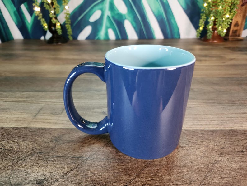 May include: A blue ceramic mug with a light blue interior. The mug has a large handle and a smooth, glossy finish. The mug is sitting on a wooden surface.
