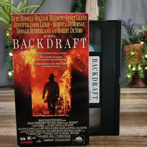 Peut inclure: Une cassette VHS et son coffret pour le film "Backdraft". La boîte présente une image enflammée d'un pompier et le titre du film en lettres grasses. La cassette est noire avec une étiquette blanche. Le coffret comprend les noms des acteurs et du réalisateur.