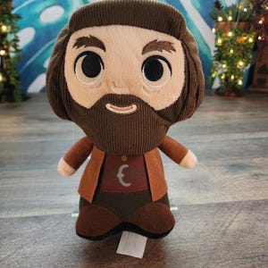 May include: A plush doll with a brown corduroy beard, hair, and clothing. The doll has a tan face with large black eyes and a stitched smile. It wears a brown jacket over a maroon shirt with a white symbol. The doll is standing on a wooden surface.