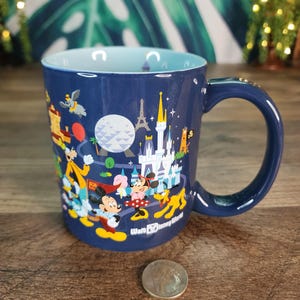May include: A navy blue ceramic mug with a light blue interior, featuring a colorful illustration of Disney characters and landmarks. The mug has a curved handle and the words "Walt Disney World" are printed on the side.