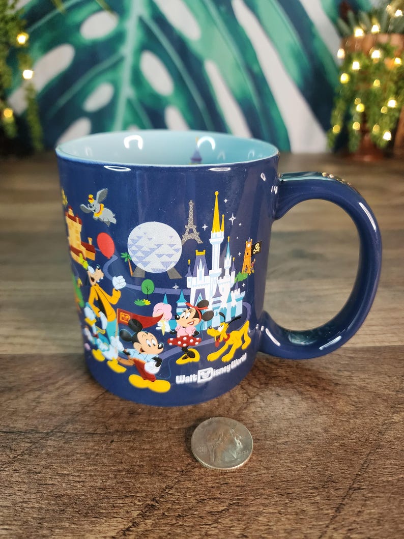 May include: A navy blue ceramic mug with a light blue interior, featuring a colorful illustration of Disney characters and landmarks. The mug has a curved handle and the words "Walt Disney World" are printed on the side.