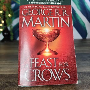 May include: A paperback book titled "A Feast for Crows" by George R.R. Martin. The cover is red with a gold chalice illustration. The book is part of the "Game of Thrones" series, a New York Times bestseller.