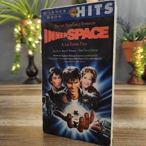 Peut inclure: Une cassette VHS Warner Bros. Hits du film L'Aventure intérieure, réalisé par Joe Dante et présenté par Steven Spielberg. La jaquette affiche le titre en grandes lettres rouges, une image centrale avec trois acteurs et une main tenant un véhicule.