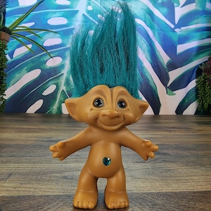 May include: A vintage Troll doll with teal hair and a gemstone belly button. The doll has a tan body, black eyes, and outstretched arms. The background features a blue and green tropical leaf pattern.