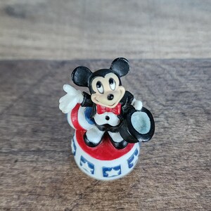 May include: A small figurine of Mickey Mouse dressed in a tuxedo, red bow tie, and top hat. He is standing on a red, white, and blue base with star designs. The figure has black ears and a smiling face.