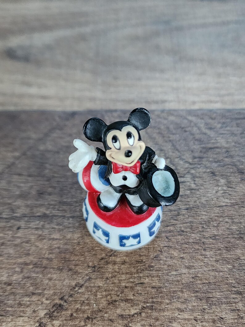 May include: A small figurine of Mickey Mouse dressed in a tuxedo, red bow tie, and top hat. He is standing on a red, white, and blue base with star designs. The figure has black ears and a smiling face.