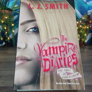 May include: A paperback book titled The Vampire Diaries: The Fury and Dark Reunion by L. J. Smith. The cover features a close-up of a person with blonde hair and a bright blue eye, with the title in red script and white text on a banner.