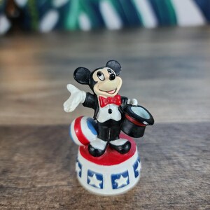 May include: A small Mickey Mouse figurine in a magician's outfit. Mickey is wearing a black tuxedo, red bow tie, and holding a black top hat. He stands on a red, white, and blue circus-themed base with star details.
