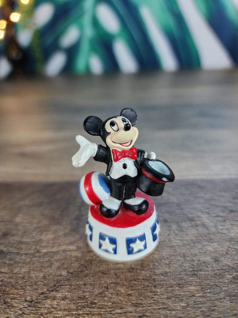 May include: A small Mickey Mouse figurine in a magician's outfit. Mickey is wearing a black tuxedo, red bow tie, and holding a black top hat. He stands on a red, white, and blue circus-themed base with star details.