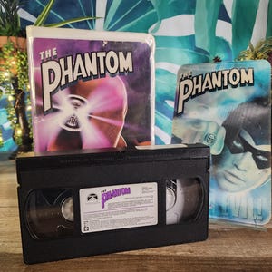 Może przedstawiać: Czarna kaseta VHS z filmem "The Phantom", z dwoma pasującymi okładkami filmowymi w tle. Okładki przedstawiają tytuł filmu w kolorze białym i fioletowo-niebieską kolorystykę. Kaseta VHS ma białą etykietę.