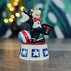 May include: A figurine of a cartoon mouse in a tuxedo, holding a top hat. The mouse stands on a base decorated with stars and stripes, with a red, white, and blue ball nearby. The figurine is a collectible item.