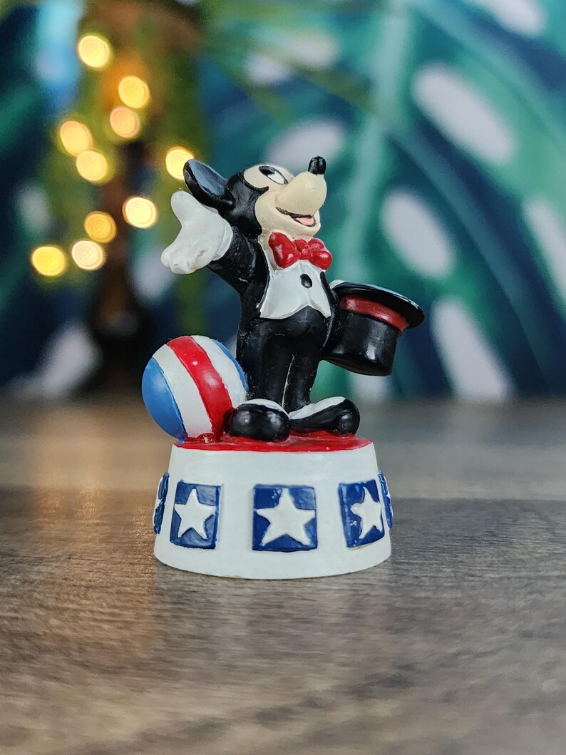 May include: A figurine of a cartoon mouse in a tuxedo, holding a top hat. The mouse stands on a base decorated with stars and stripes, with a red, white, and blue ball nearby. The figurine is a collectible item.