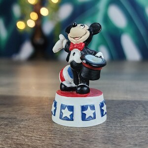 May include: A figurine of Mickey Mouse dressed as a magician, wearing a tuxedo and a red bow tie. He is standing on a red and white platform with blue star accents. The figurine is a collectible item.