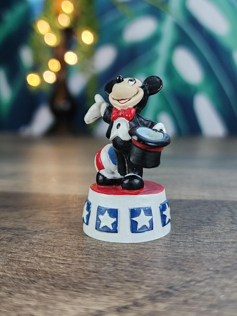 May include: A figurine of Mickey Mouse dressed as a magician, wearing a tuxedo and a red bow tie. He is standing on a red and white platform with blue star accents. The figurine is a collectible item.