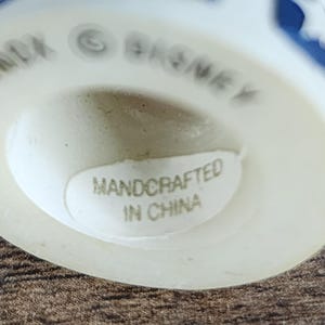 May include: A white ceramic figurine with blue star-shaped designs. The base of the figurine has the text "HANDCRAFTED IN CHINA" on a sticker. The word "DISNEY" is also visible.