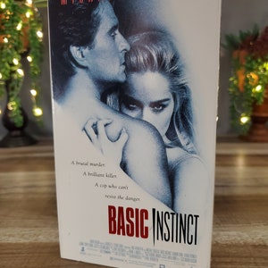 May include: A VHS tape for the movie "Basic Instinct" featuring Michael Douglas and Sharon Stone. The cover art shows a man and woman in a close embrace, with the title in red. Text on the cover reads "A brutal murder. A brilliant killer. A cop who can't resist the danger."