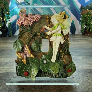 May include: A decorative light switch cover featuring a fairy with blonde hair and wings, dressed in white and green. The cover has a garden scene with butterflies, flowers, and ladybugs. The switch plate is made of clear acrylic.