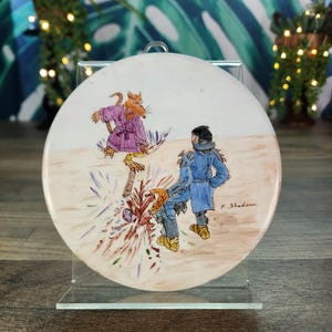 May include: A round, off-white decorative plate featuring a whimsical illustration. The artwork depicts a cat in a purple robe, a figure in blue, and a burst of color. The plate is displayed on a clear stand.