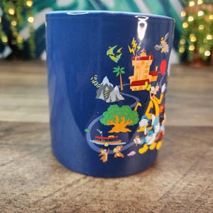 May include: A cylindrical, cobalt blue ceramic mug featuring a colorful cartoon illustration. The design includes a castle, mountains, a tree, and various cartoon characters. The mug is likely a souvenir or collectible item.