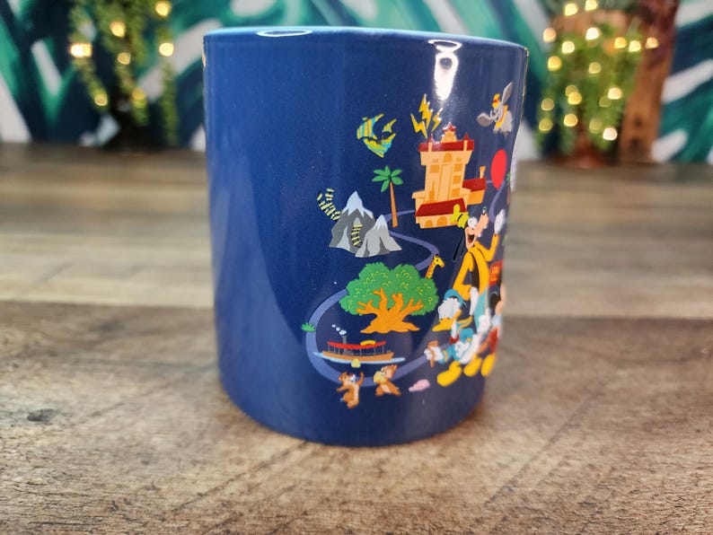 May include: A cylindrical, cobalt blue ceramic mug featuring a colorful cartoon illustration. The design includes a castle, mountains, a tree, and various cartoon characters. The mug is likely a souvenir or collectible item.