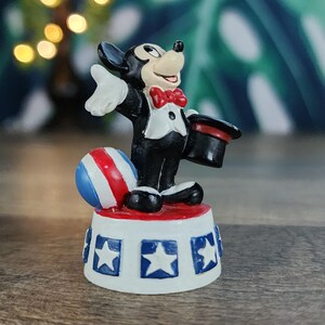 May include: A figurine of a cartoon mouse in a tuxedo, holding a top hat and standing on a platform with stars. A red, white, and blue ball is next to the mouse. The figurine is a collectible item.