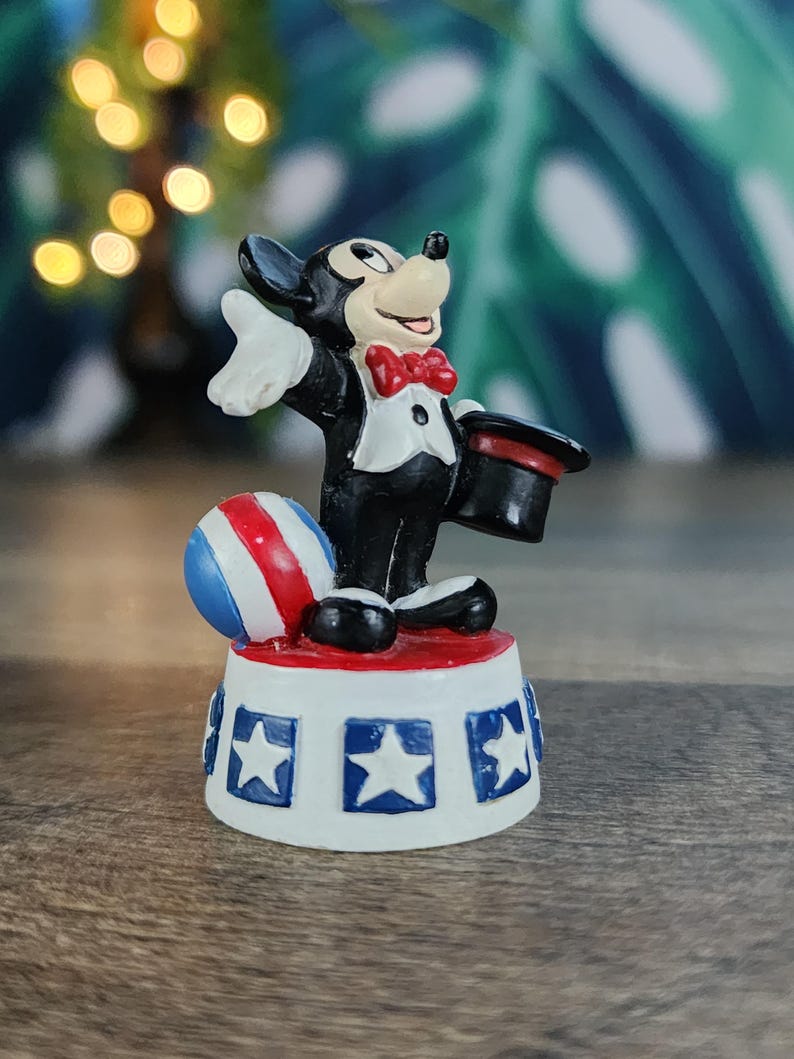 May include: A figurine of a cartoon mouse in a tuxedo, holding a top hat and standing on a platform with stars. A red, white, and blue ball is next to the mouse. The figurine is a collectible item.