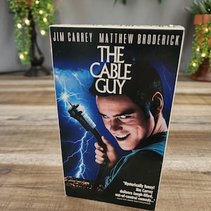 May include: A VHS tape for the movie "The Cable Guy" featuring Jim Carrey and Matthew Broderick. The cover art shows Jim Carrey holding a cable tool with a lightning bolt background. The text on the cover reads "The Cable Guy".