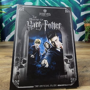 Puede incluir: Un libro de tapa dura titulado "The Making of Harry Potter: The Official Guide". La portada oscura presenta imágenes de los personajes de Harry Potter y el logotipo de Warner Bros. Studio Tour.
