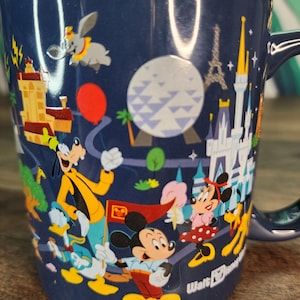 May include: Navy blue ceramic mug featuring Disney characters and landmarks. The mug showcases Mickey Mouse, Minnie Mouse, Goofy, Donald Duck, and Dumbo, along with the Eiffel Tower and Cinderella's castle. The mug has a dark blue handle and the Walt Disney logo.