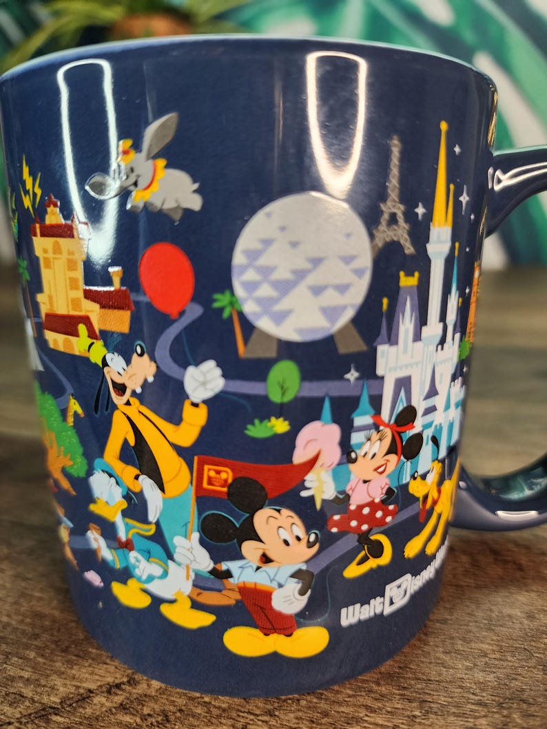 May include: Navy blue ceramic mug featuring Disney characters and landmarks. The mug showcases Mickey Mouse, Minnie Mouse, Goofy, Donald Duck, and Dumbo, along with the Eiffel Tower and Cinderella's castle. The mug has a dark blue handle and the Walt Disney logo.