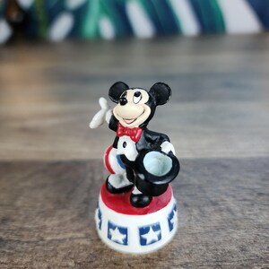 May include: A Mickey Mouse figurine in a classic outfit, holding a top hat and a white glove. The figure stands on a red and white base with blue star accents. The figurine is a collectible item.