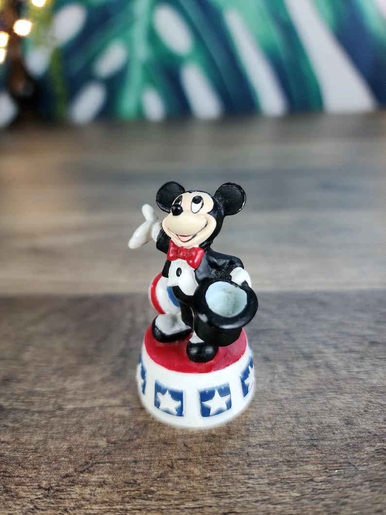 May include: A Mickey Mouse figurine in a classic outfit, holding a top hat and a white glove. The figure stands on a red and white base with blue star accents. The figurine is a collectible item.