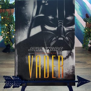 Puede incluir: Un libro de tapa dura titulado "Star Wars: The Complete Vader" con una imagen en blanco y negro del casco de Darth Vader. El libro se muestra en un soporte en forma de flecha azul oscuro.