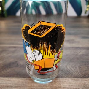 Puede incluir: Un vaso transparente con un gráfico de dibujos animados de una caja que explota. La caja es amarilla y negra, con una cinta roja. El vaso tiene una base azul claro y está sobre una superficie de madera.