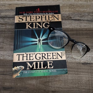 May include: A copy of Stephen King's "The Green Mile" with black-framed glasses resting on the cover. The book is a New York Times bestseller, with the title and author's name prominently displayed. The cover features a green and black design.