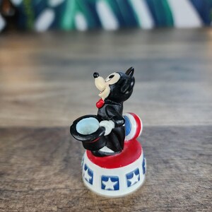 May include: A small, vintage figurine of a black cat in a magician's outfit, complete with a top hat and red bow tie. The cat is perched on a red, white, and blue circus-style base with star details. A whimsical collectible.