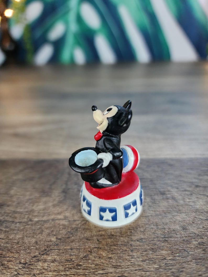 May include: A small, vintage figurine of a black cat in a magician's outfit, complete with a top hat and red bow tie. The cat is perched on a red, white, and blue circus-style base with star details. A whimsical collectible.