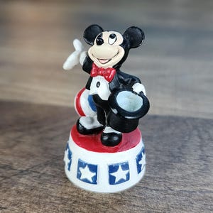 May include: A small figurine of Mickey Mouse in a tuxedo, holding a top hat and a white glove. He stands on a red, white, and blue base with star designs. The figure is black, white, red, and blue.