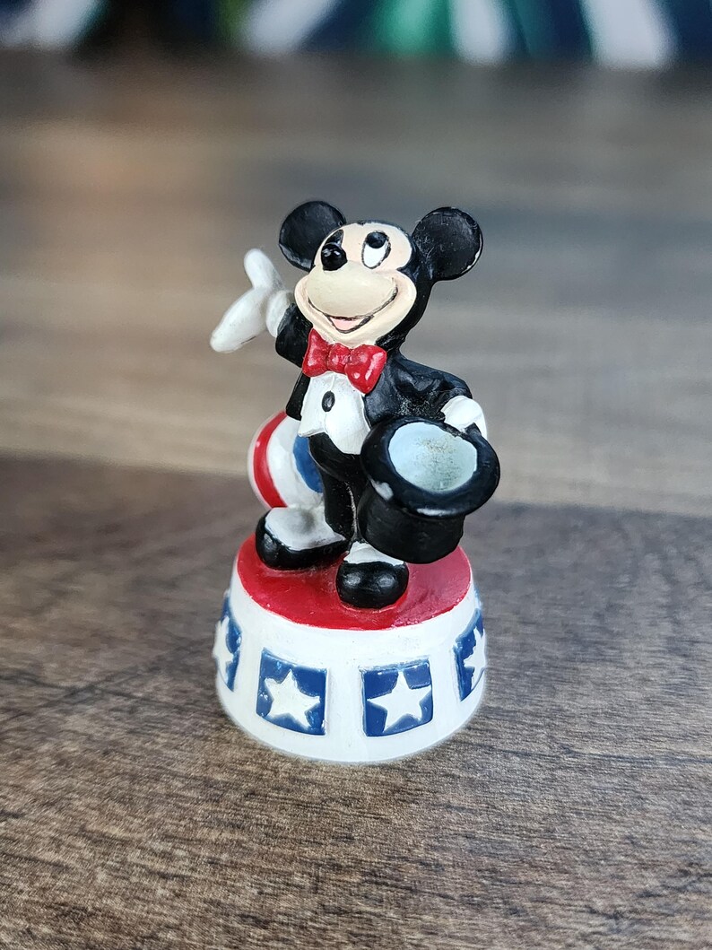 May include: A small figurine of Mickey Mouse in a tuxedo, holding a top hat and a white glove. He stands on a red, white, and blue base with star designs. The figure is black, white, red, and blue.